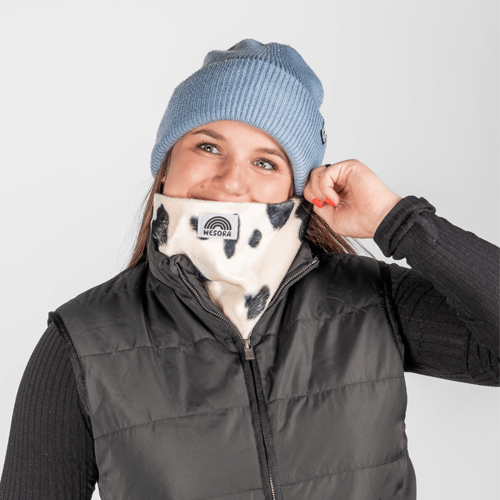 Printed Neckwarmer - Cow Spirit Fur mit Kuhmuster und WESORA-Logo, getragen von jungem Erwachsenen, der Gaiter zum Gesicht zieht, vor neutralem Hintergrund.