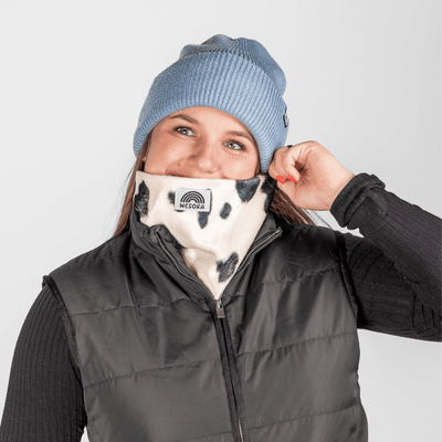 Printed Neckwarmer - Cow Spirit Fur mit Kuhmuster und WESORA-Logo, getragen von jungem Erwachsenen, der Gaiter zum Gesicht zieht, vor neutralem Hintergrund.