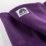 Fleece Neckwarmer - Purple Noir mit WESORA-Label und Regenbogengrafik, auf einem Tisch liegend, zeigt die handgemachte Qualität und Eleganz des Accessoires.