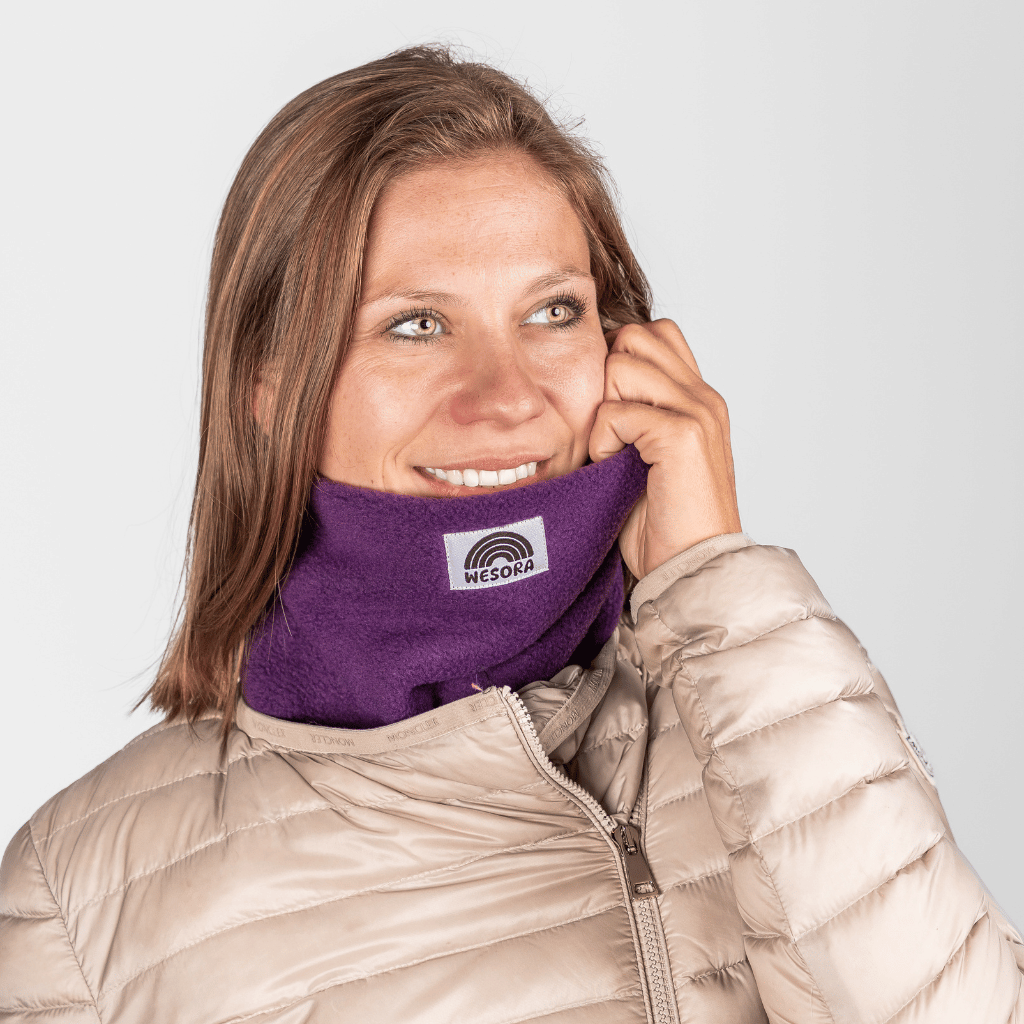 Fleece Neckwarmer - Purple Noir mit Regenbogenlogo, getragen von lächelnder Frau, die den Rand anpasst, fotografiert vor grauem Hintergrund.