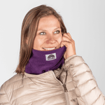 Fleece Neckwarmer - Purple Noir mit Regenbogenlogo, getragen von lächelnder Frau, die den Rand anpasst, fotografiert vor grauem Hintergrund.