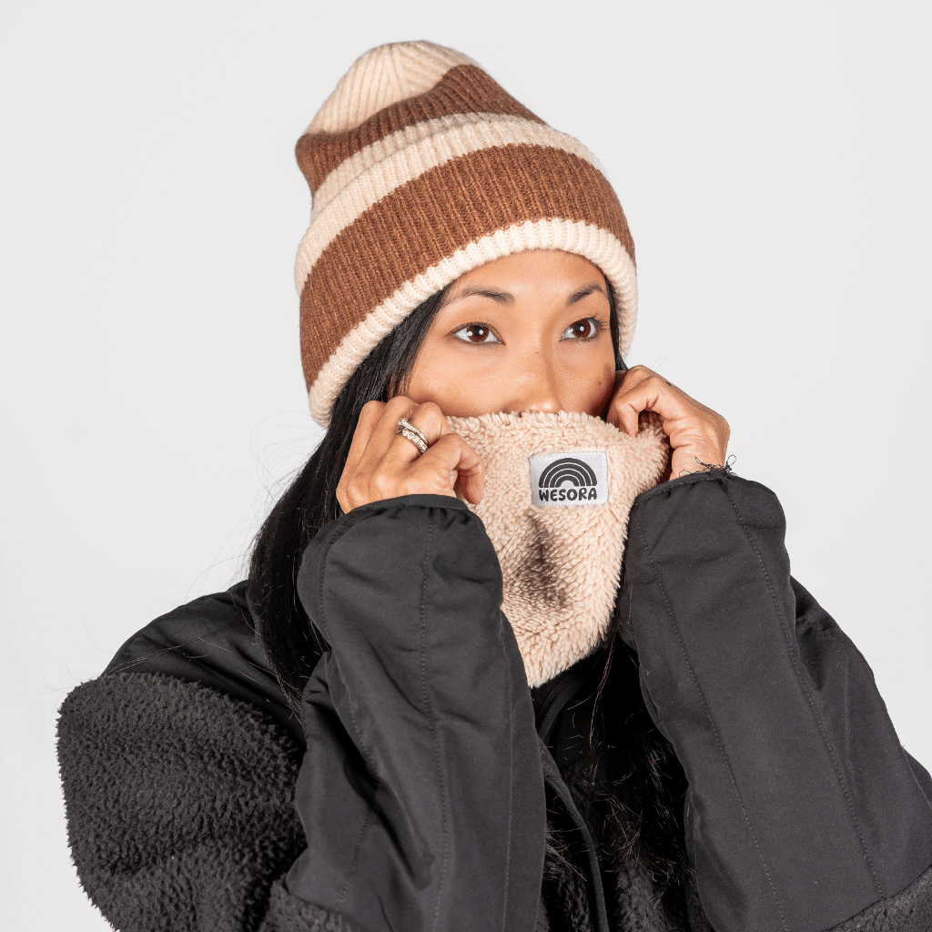 Sherpa Neckwarmer - Fluffy Beige, beige aus Sherpa-Fleece, getragen von Person in schwarzer Jacke, zieht hoch bis unter die Augen, in Studioaufnahme.