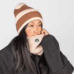 Sherpa Neckwarmer - Fluffy Beige, getragen von Person mit gestreifter Mütze und dunklem Haar, hält den weichen, beigen Fleece-Nackenwärmer über Gesicht.