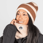 Sherpa Neckwarmer - Fluffy Beige, getragen von einer lächelnden Frau mit brauner Strickmütze, fotografiert vor einem grauen Hintergrund, von vorn leicht seitlich.