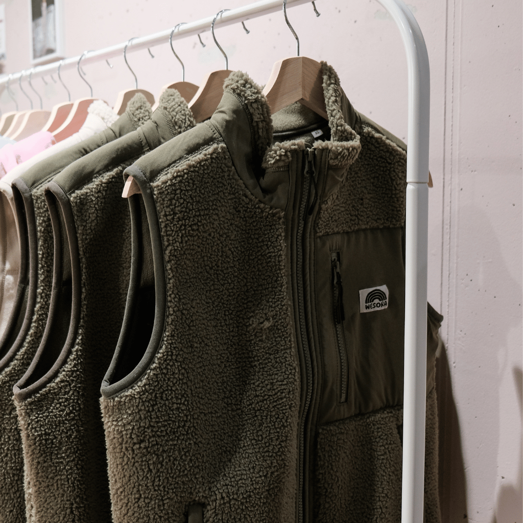 Sherpa Weste - Unisex mit Reißverschlussdesign und hoher Kragen, präsentiert auf Holzkleiderbügeln an einem weißen Kleiderständer.