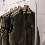 Sherpa Weste - Unisex mit Reißverschlussdesign und hoher Kragen, präsentiert auf Holzkleiderbügeln an einem weißen Kleiderständer.