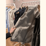 Sherpa Weste - Unisex, cremefarben, mit Reißverschluss, gehalten von Person, vor einer Auswahl an dunkleren Westen und Langarmshirts in einem Laden.