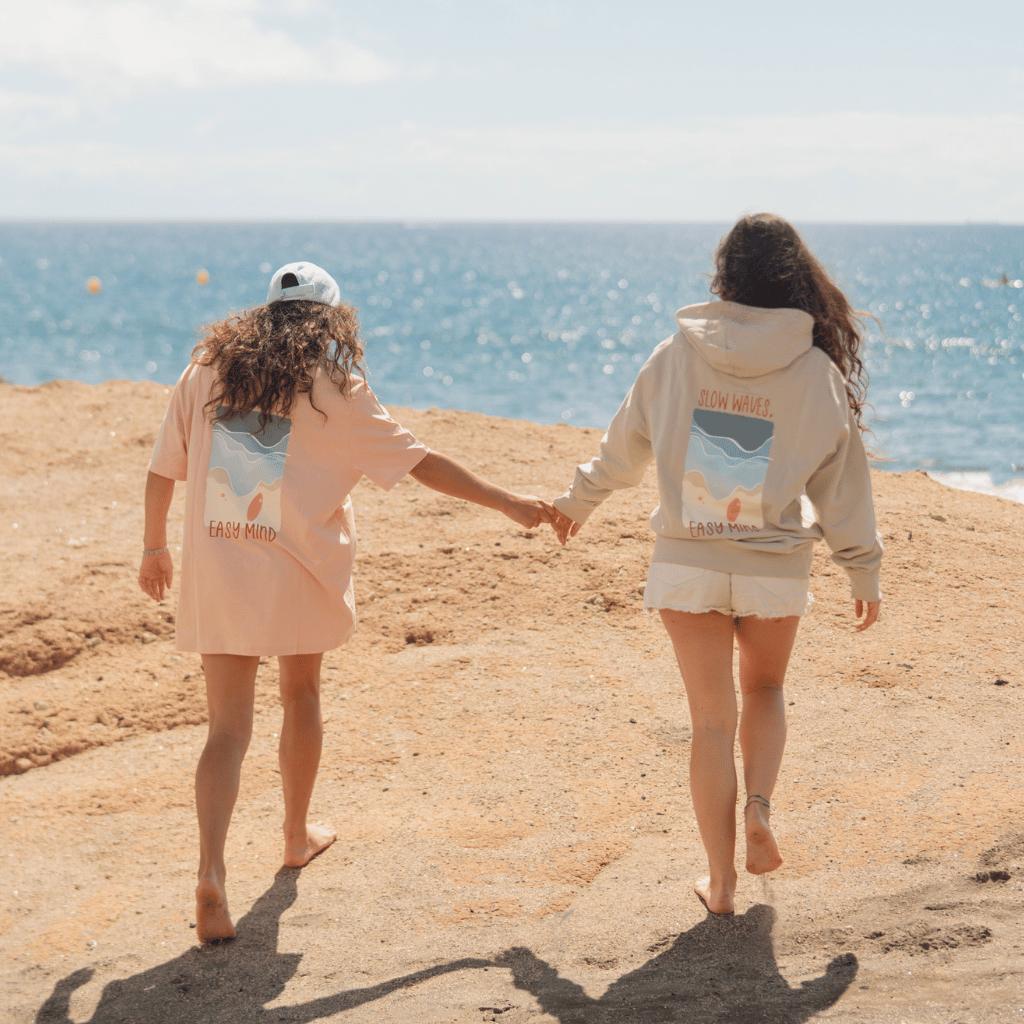 Slow Waves - Unisex Pullover mit Rücken- und Brustprint, getragen von Paaren am Strand, händchenhaltend mit Blick zum Meer, eingefangen in ruhiger, natürlicher Atmosphäre.