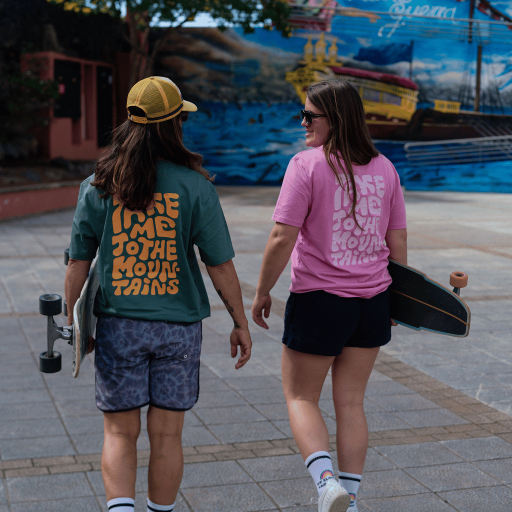 Take me to the Mountains - Unisex T-Shirt mit großem Rückendruck, getragen von zwei Personen auf einem gepflasterten Platz, beide mit Longboards in der Hand.