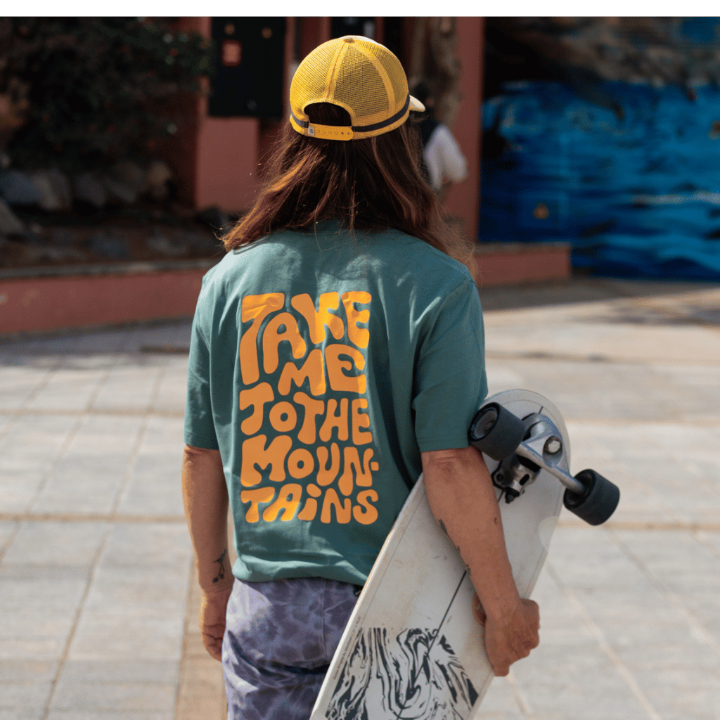 Take me to the Mountains - Unisex T-Shirt mit großem Rückenprint, getragen von Person mit Skateboard, fotografiert im Freien auf einem Platz von hinten.