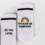 Socken - WESORA: Weiße Tennissocken mit schwarzem Schriftzug „SKI YOU LATER“ und Regenbogenmotiv „WE SLIDE ON RAINBOWS“, flach auf weißem Hintergrund.