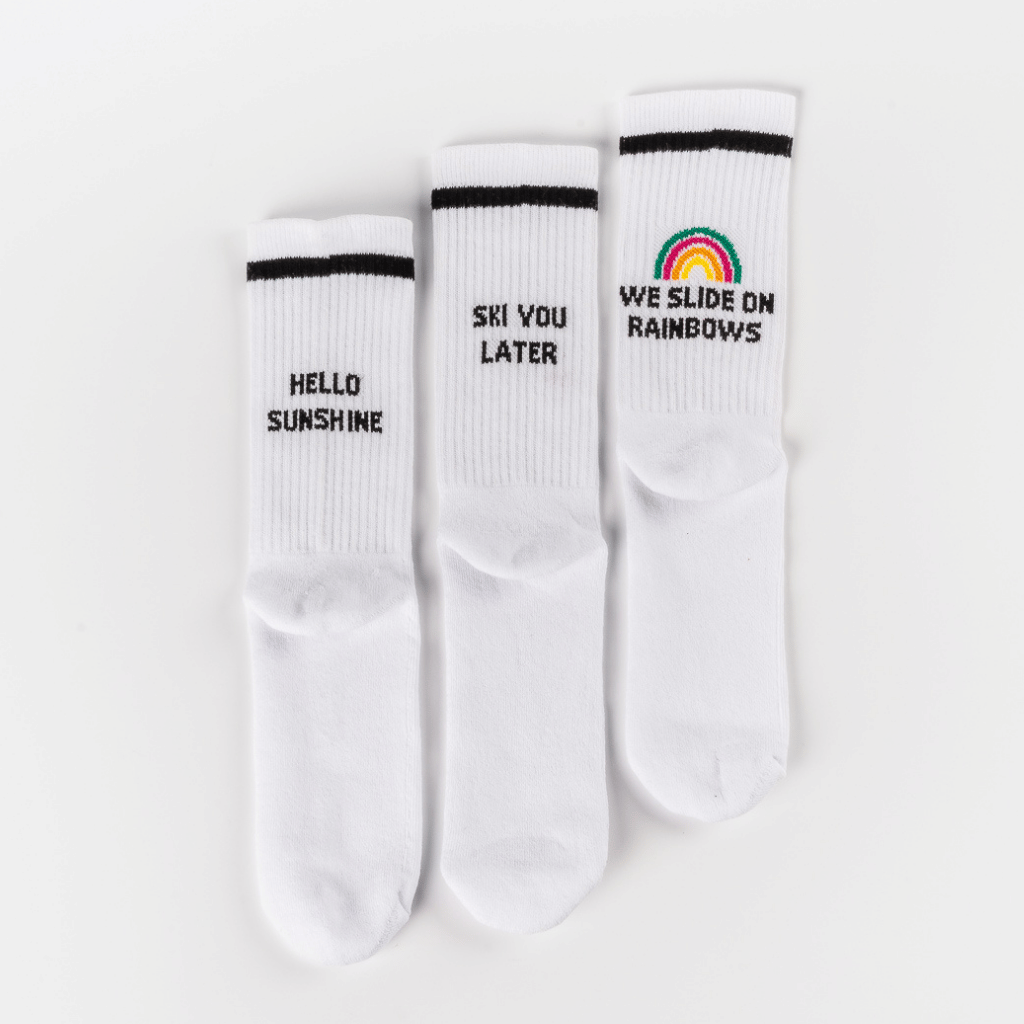 Socken-Bundle mit drei Designs: HELLO SUNSHINE, SKI YOU LATER und WE SLIDE ON RAINBOWS mit Regenbogenmotiv, flach nebeneinander liegend.