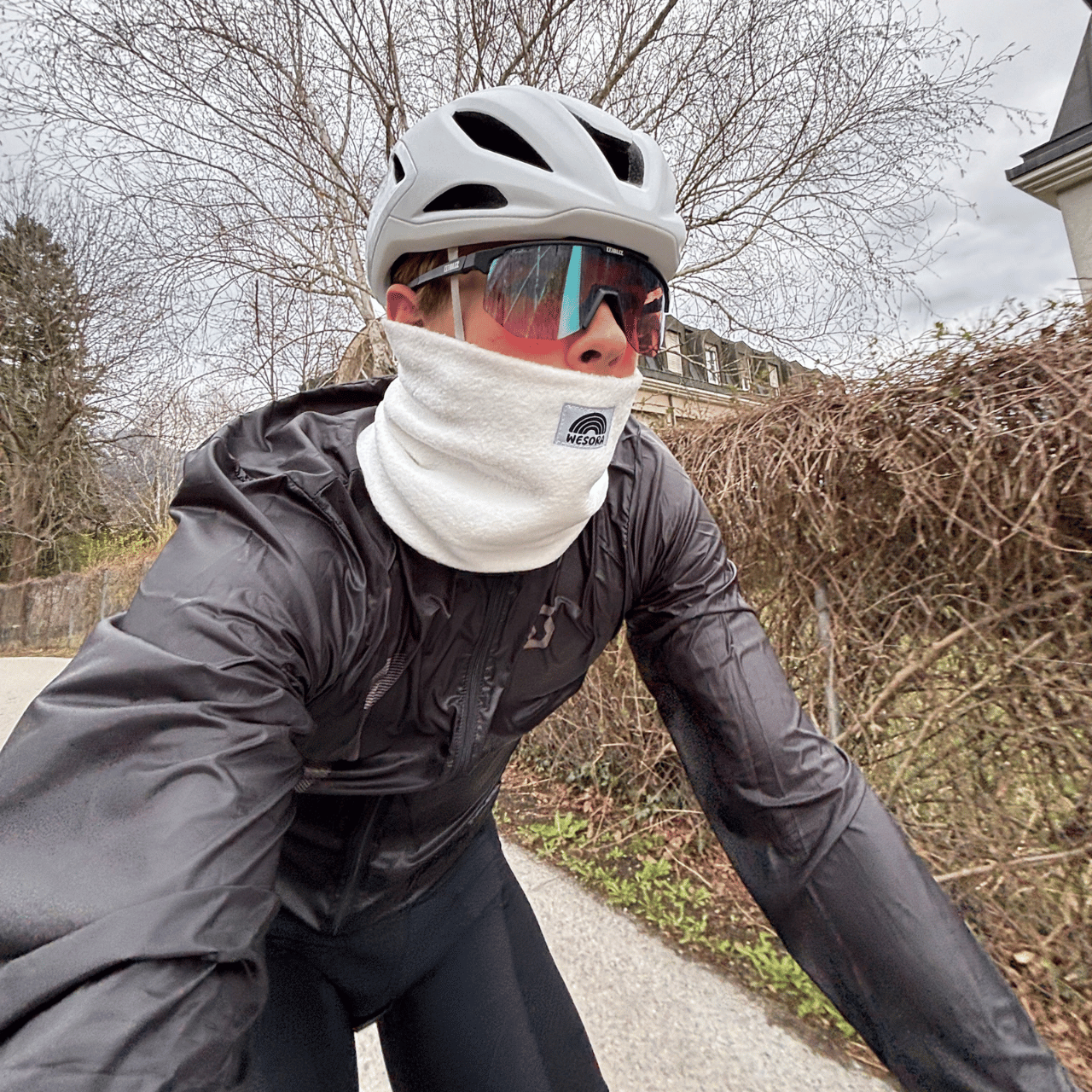 Fleece Neckwarmer - Off White, getragen von einem Radfahrer auf einer ruhigen Straße bei kühlem Wetter, mit weißem Helm und Sonnenbrille.