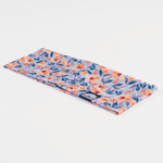 Stirnband - Wildflower mit dichtem Blumenmuster in Orange, Rot und Lila auf hellblauem Grund, flachgelegt, mit Markenlabel am Rand.