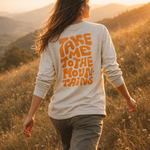 Take me to the Mountains - Unisex Longsleeve mit großem Rückendruck, getragen von Person auf grasbewachsenem Hügel bei Sonnenuntergang, von hinten gesehen.